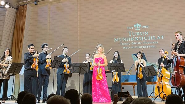 Anne-Sophie Mutter stands with her chamber ensemble on stage, wearing a pink evening gown.